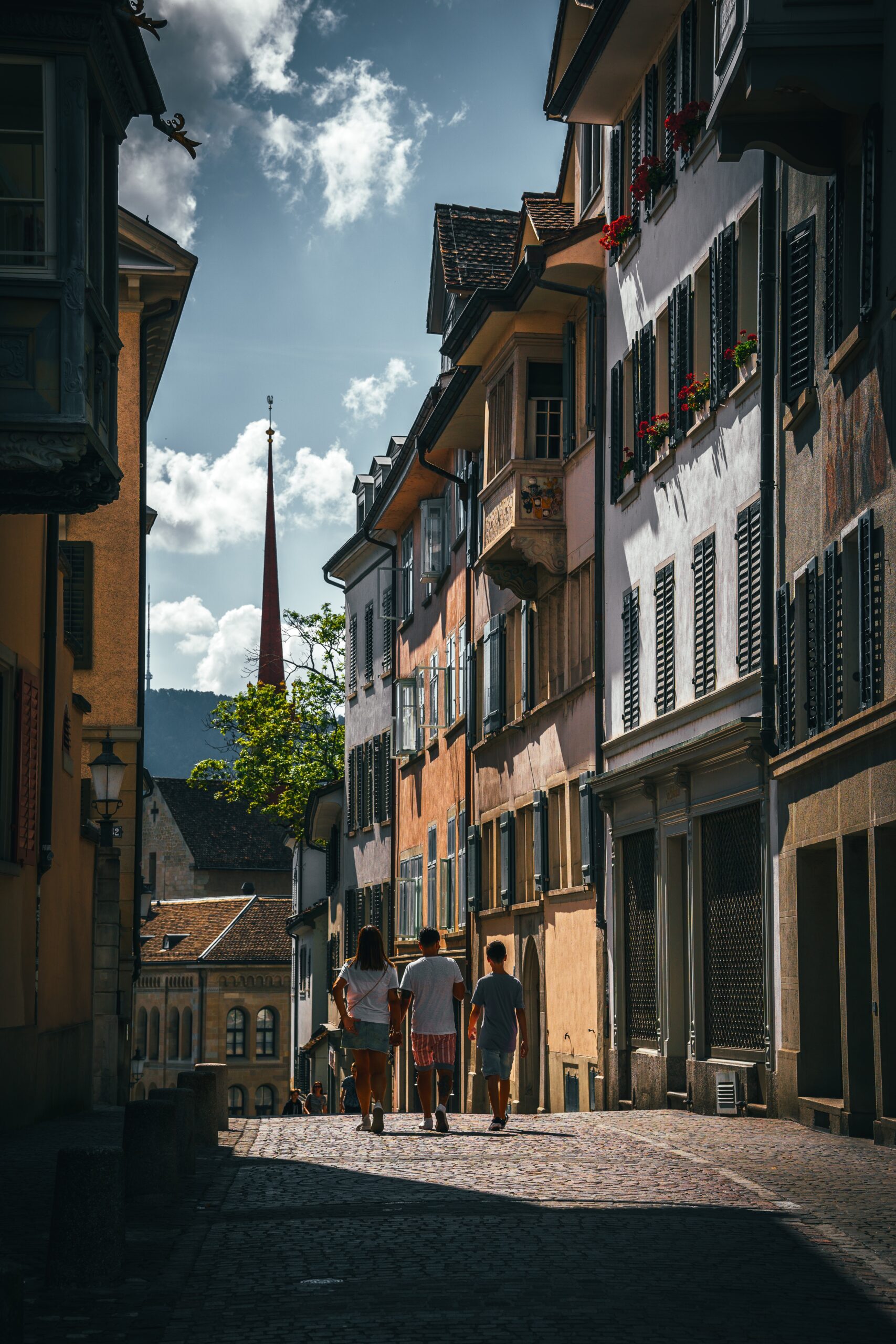 A group of people walking down a narrow street with shops and cafes on either side. The street is lined with old buildings with colorful facades