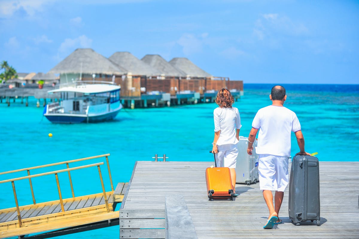 Couple rolling carry-on luggage along a dock with turquoise water and overwater bungalows