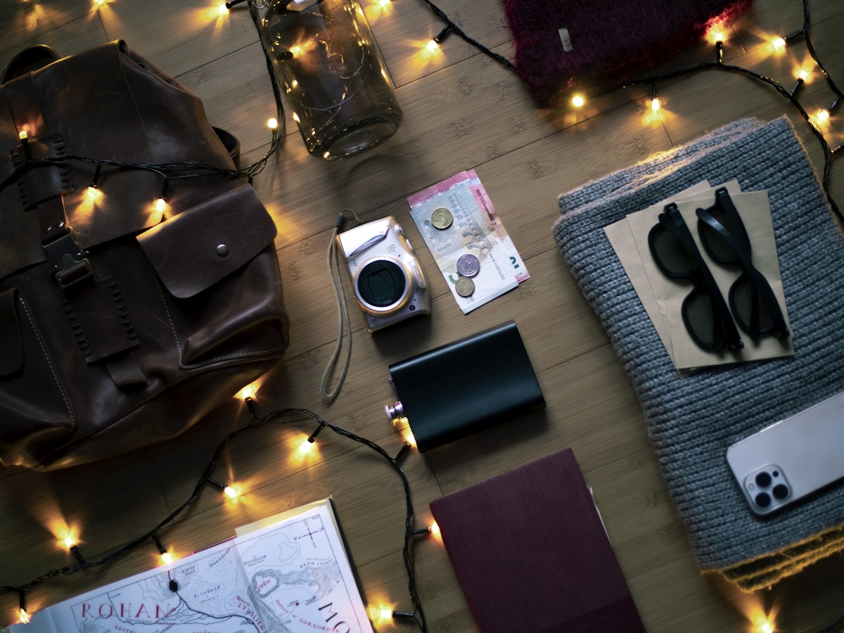Travel essentials laid out next to a leather mini backpack including camera, phone, sunglasses, passport, and portable charger
