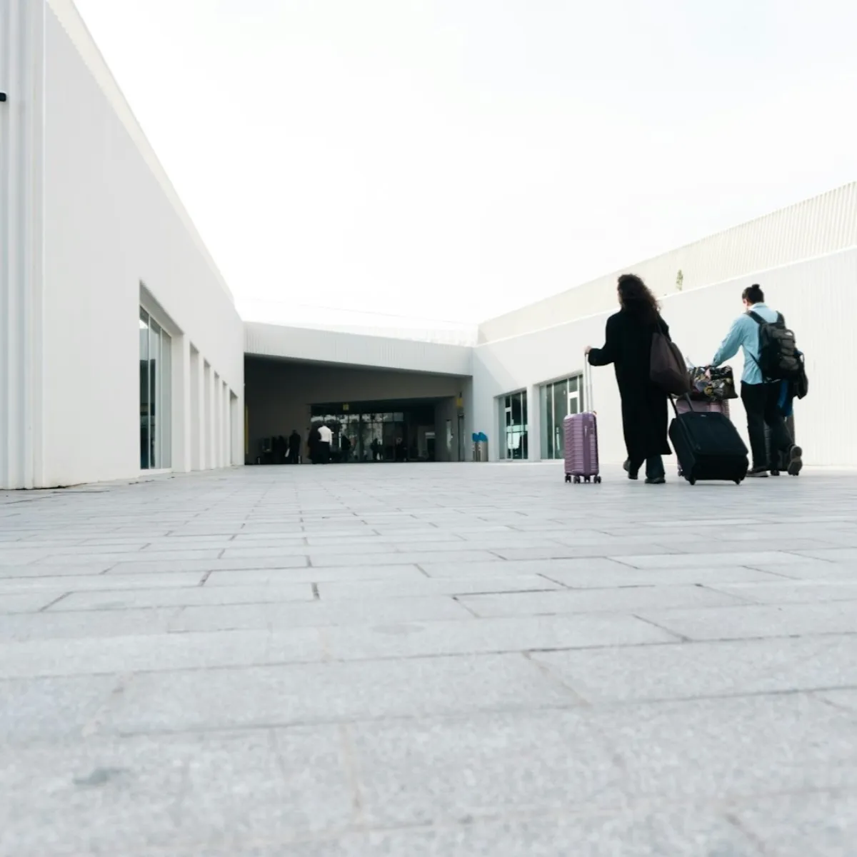 Two travelers walking toward a modern airport terminal each pulling a rolling carry-on suitcase and wearing a backpack