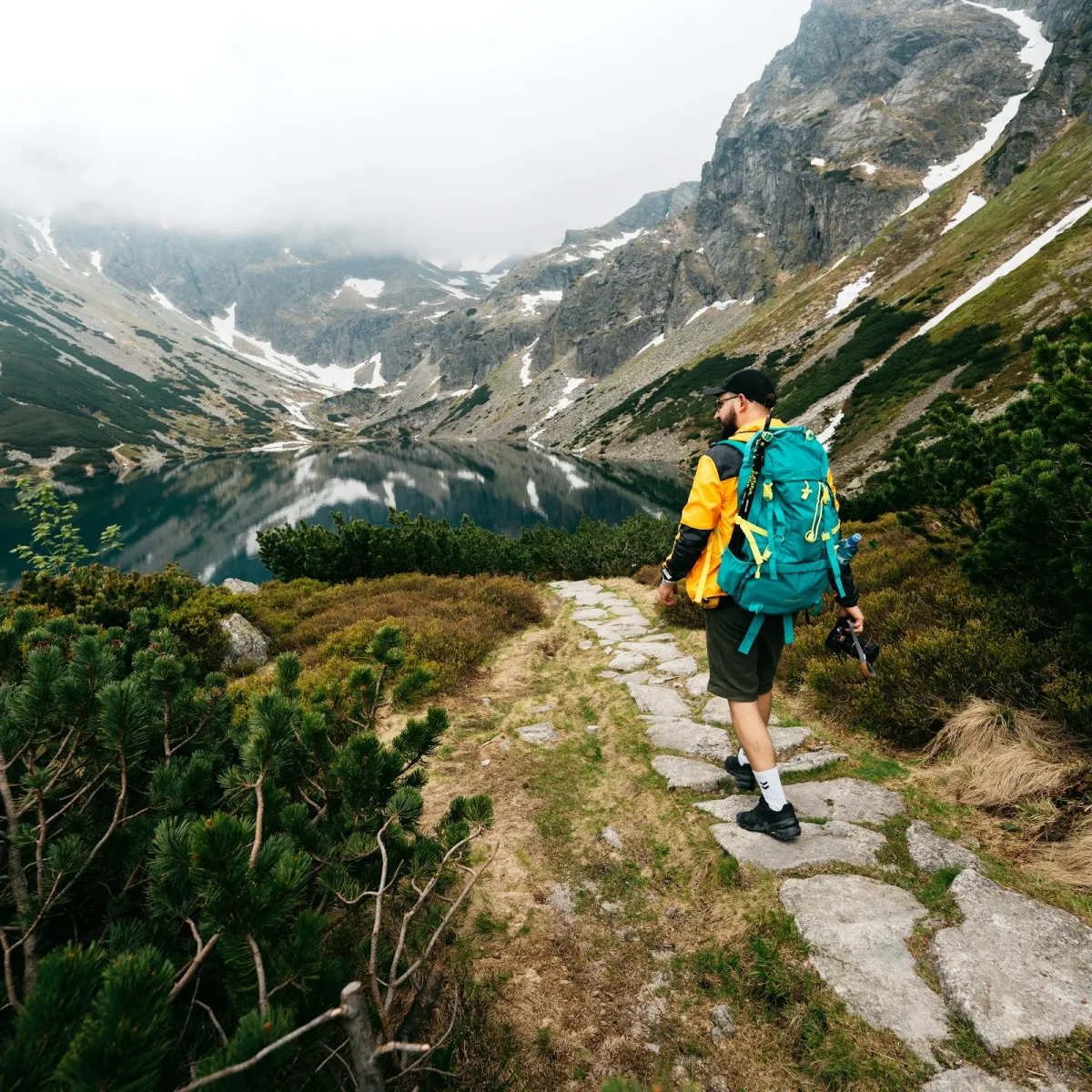 Hiker carrying a teal ultralight backpack on a rocky alpine trail overlooking a mountain lake.