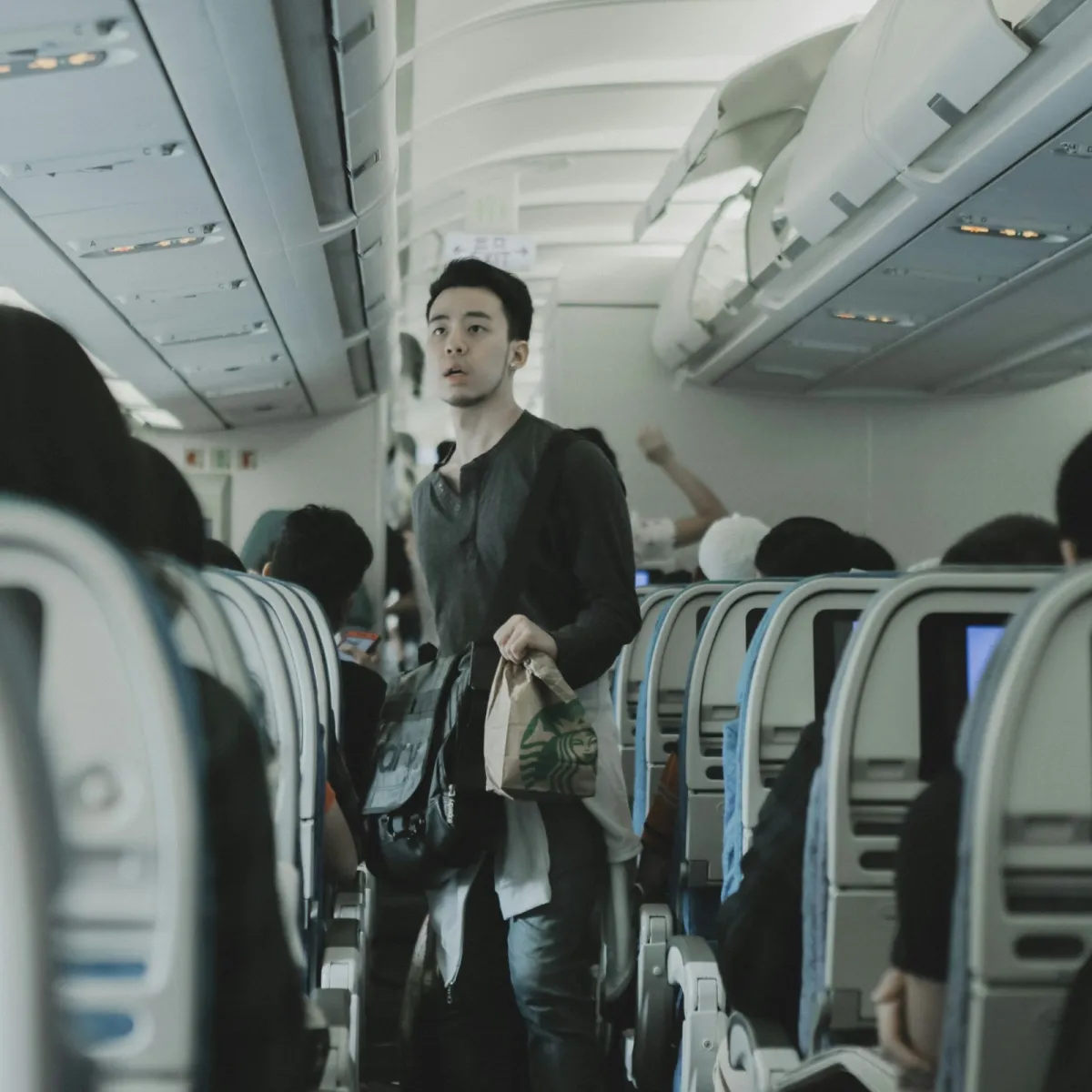 Passenger inside an airplane cabin holding a carry-on and personal item bag before stowing them at the seat