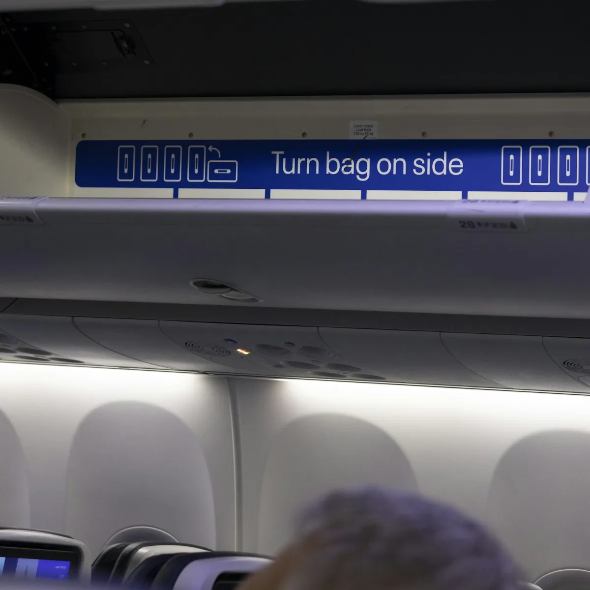 Airplane overhead bin showing the turn-bag-on-side instruction decal above passenger seats