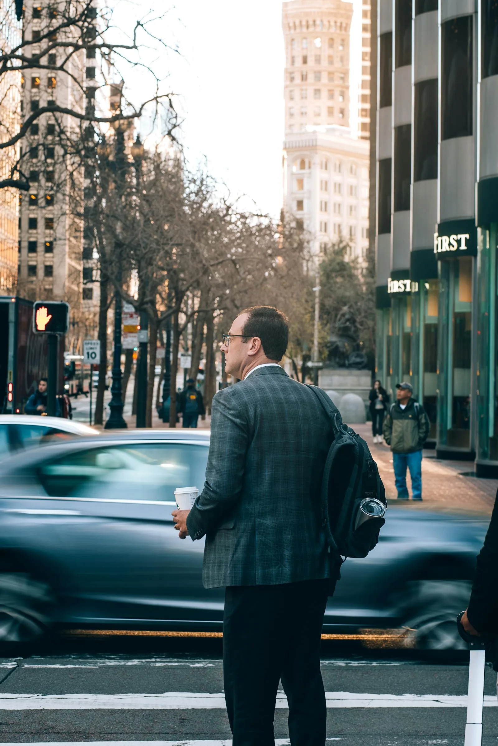 A man in a suit with a backpack waits to cross a bustling city street.