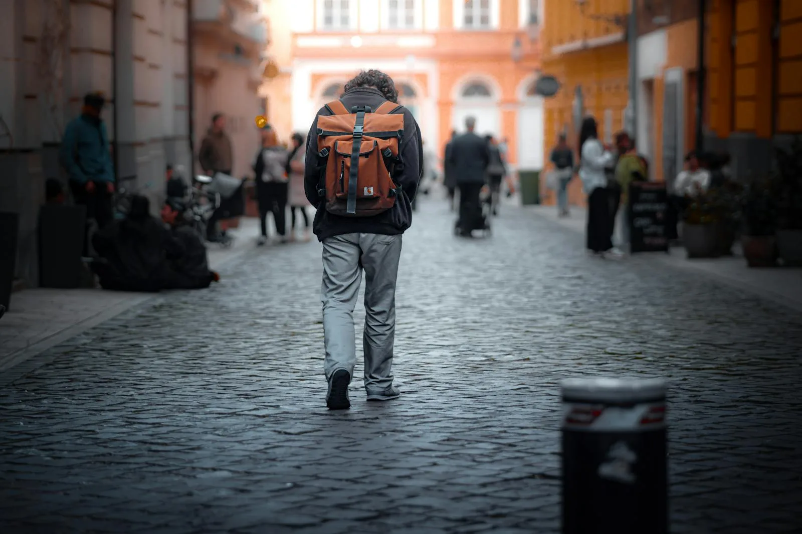 A solitary man walks down a cobblestone street with a backpack, surrounded by people in the city.