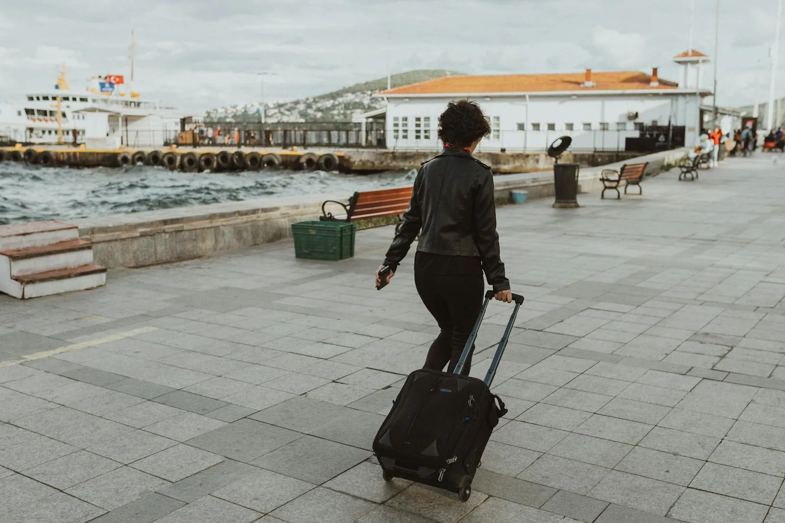A woman walks on a seaside promenade with a suitcase, capturing urban travel vibes