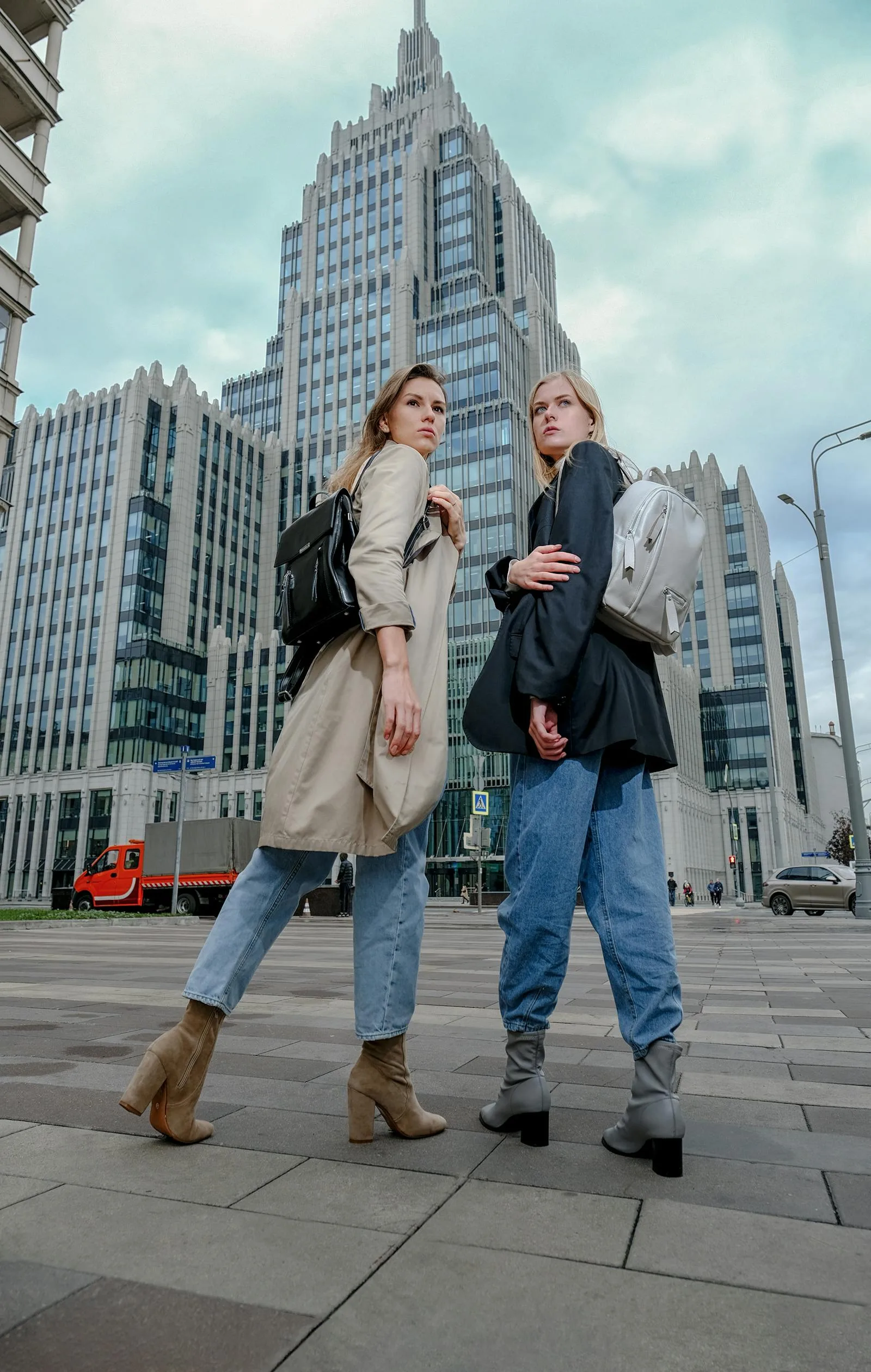 Two fashionable women with backpacks pose in a modern city setting.