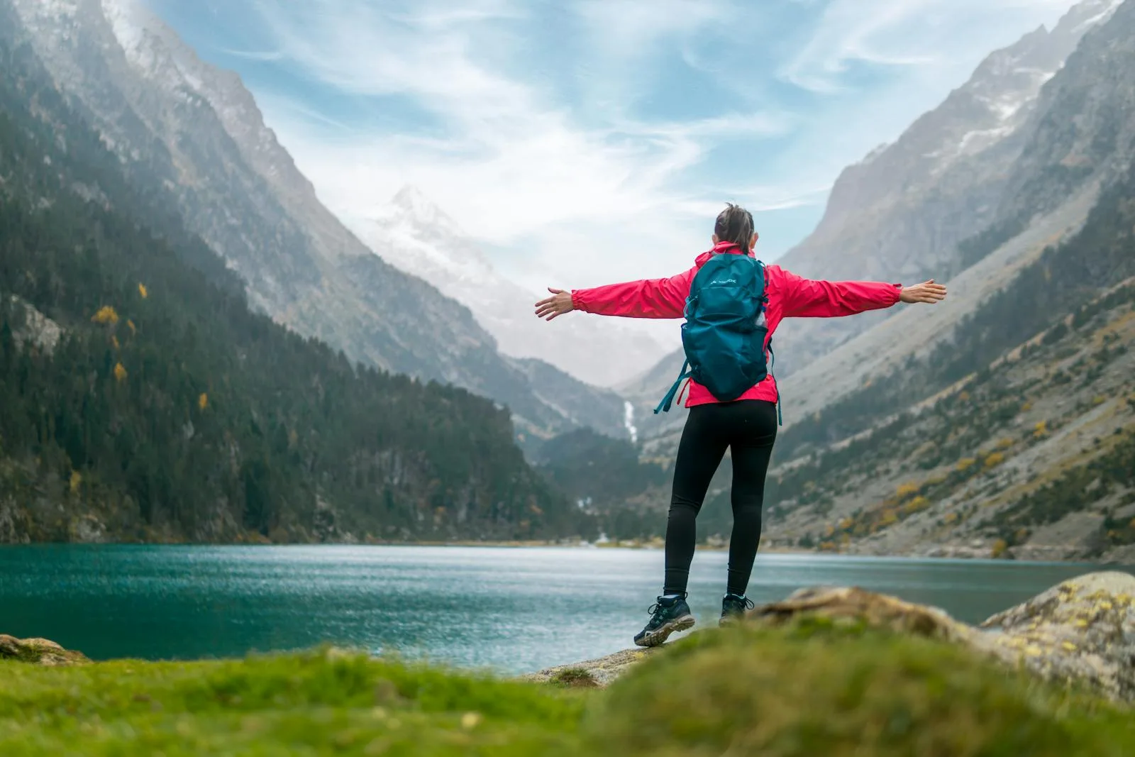 A female hiker in a red jacket stands with arms wide open, overlooking a tranquil alpine lake.