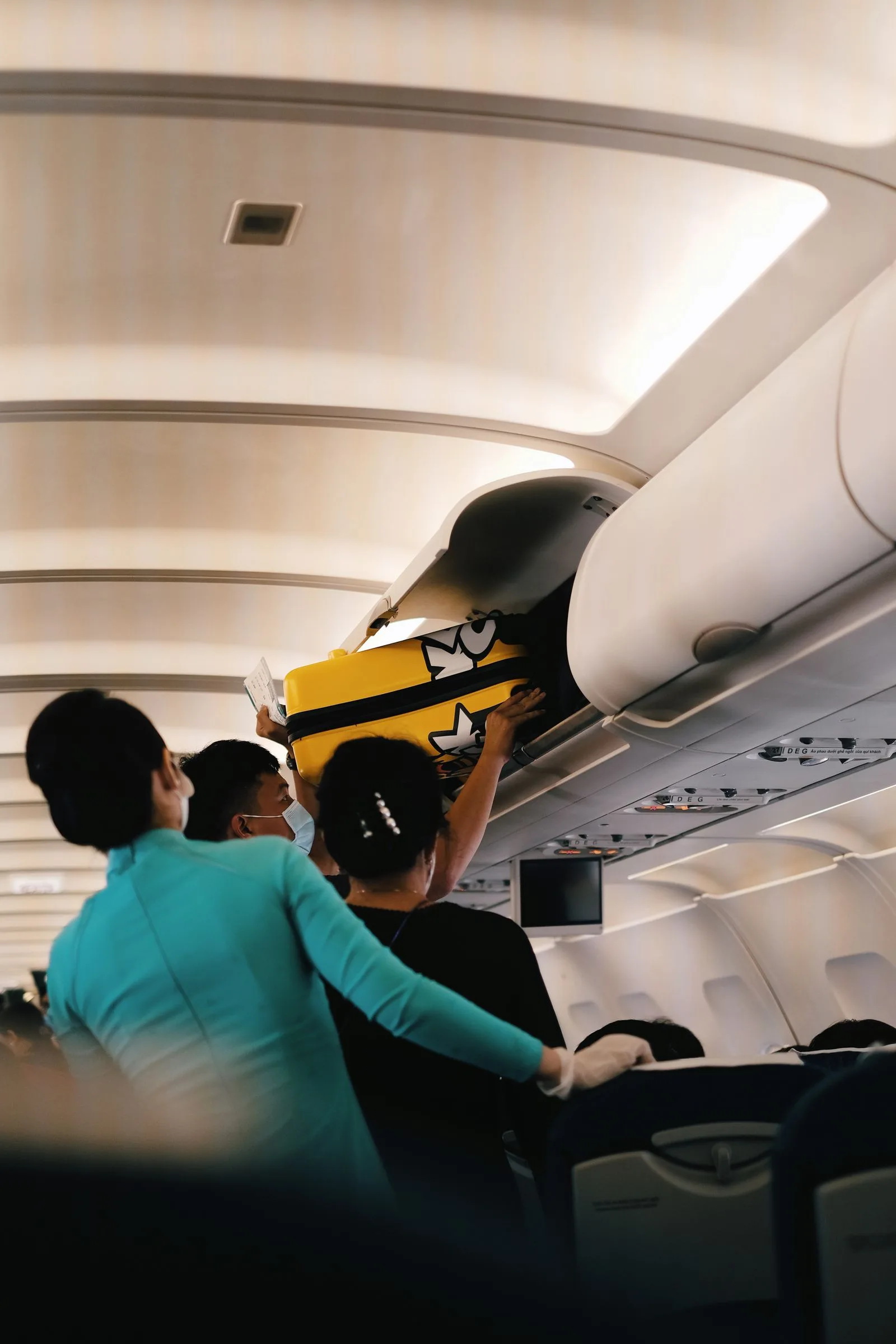 Passengers placing a yellow suitcase in the overhead bin of an airplane cabin, with crew assistance