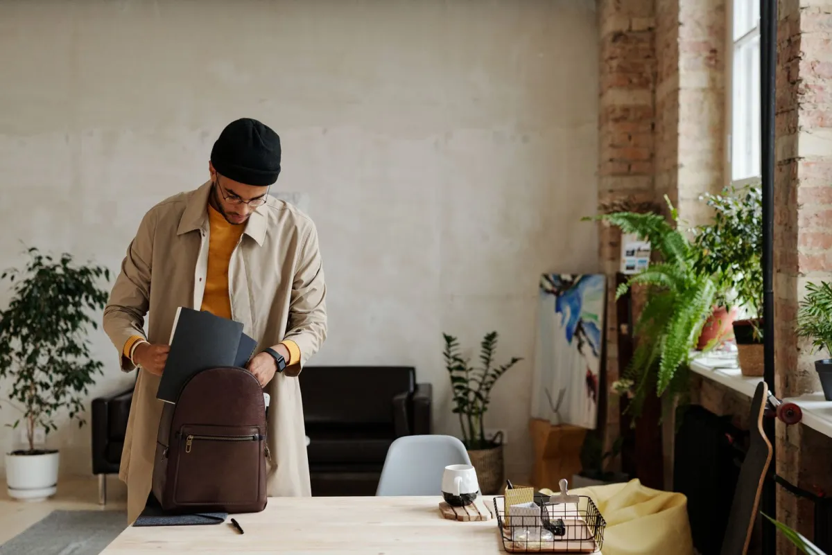 Man sliding a laptop into a backpack on a desk in a plant filled loft workspace