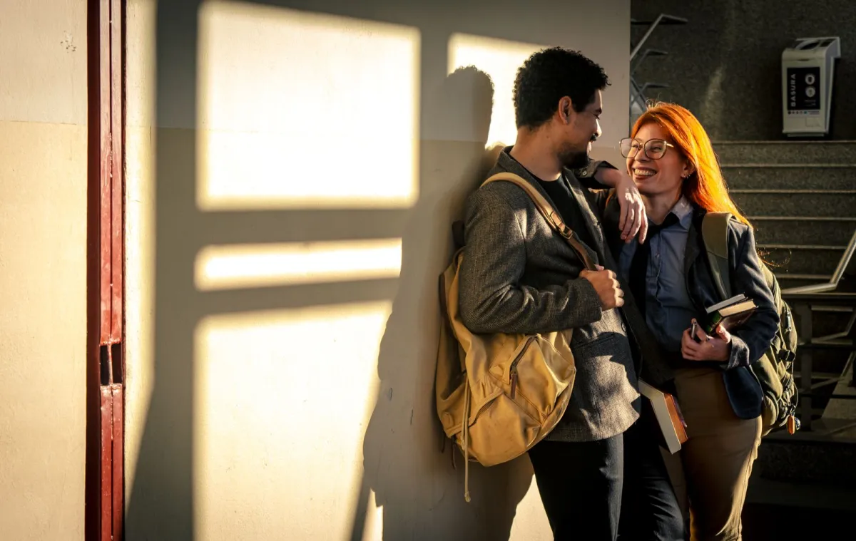 Two students carrying backpacks pause to talk in a sunlit school corridor