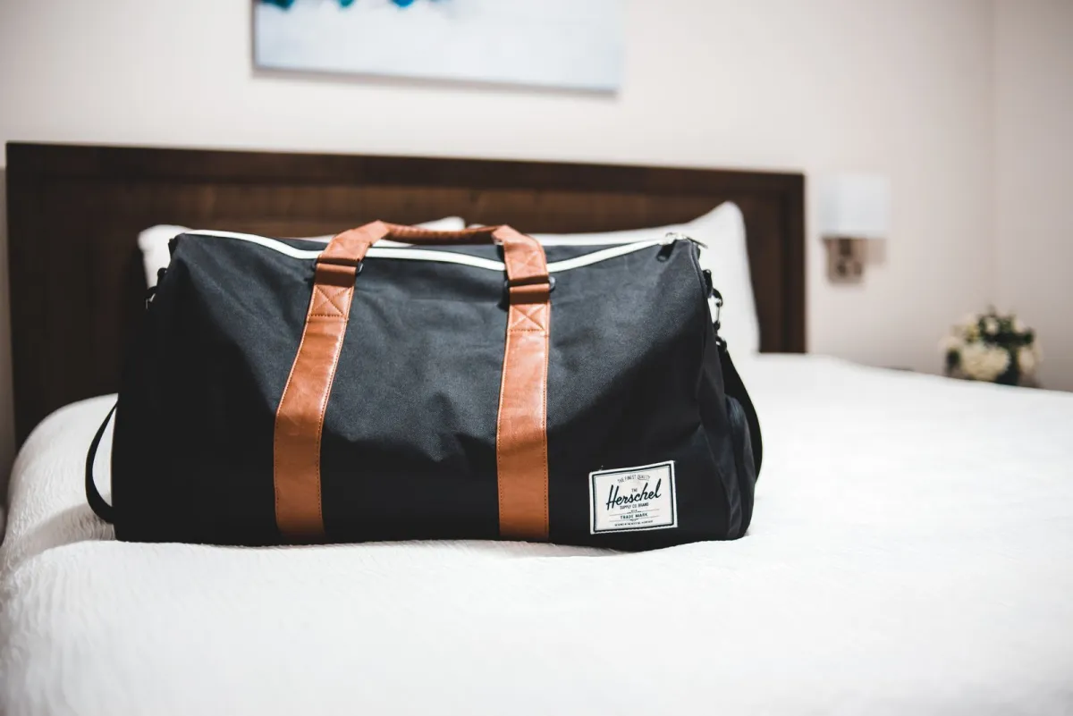 Duffel bag resting on a made hotel bed with plump white pillows and a bedside lamp