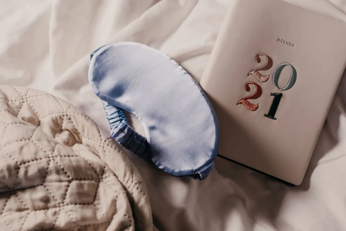 Padded satin sleep mask lying on a quilted bedspread next to a hardback planner