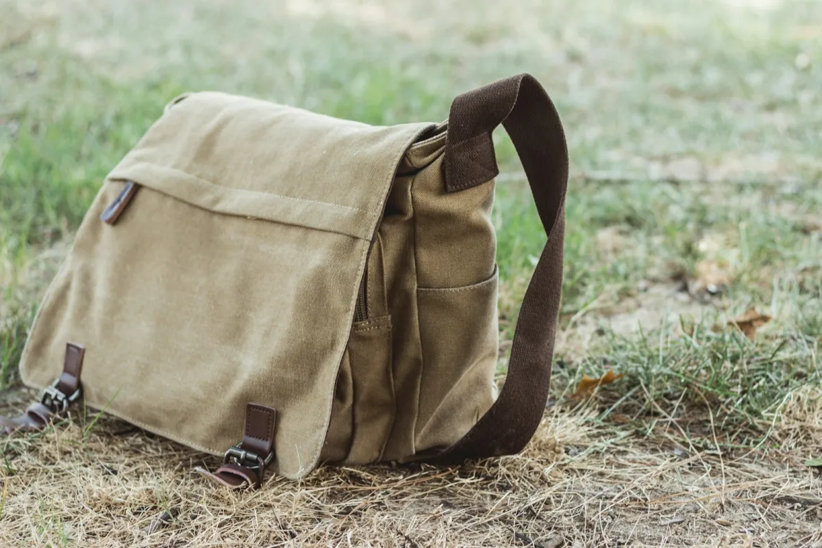 Compact tan canvas messenger bag with buckled front pockets sitting on dry grass
