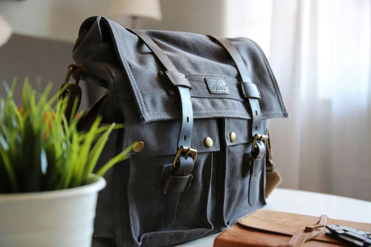Waxed canvas messenger bag with leather buckle straps on a table beside a potted plant