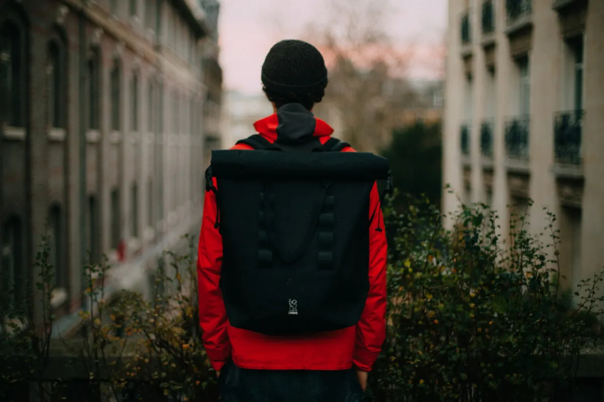 Commuter in a red jacket walking a city street wearing a black roll-top backpack