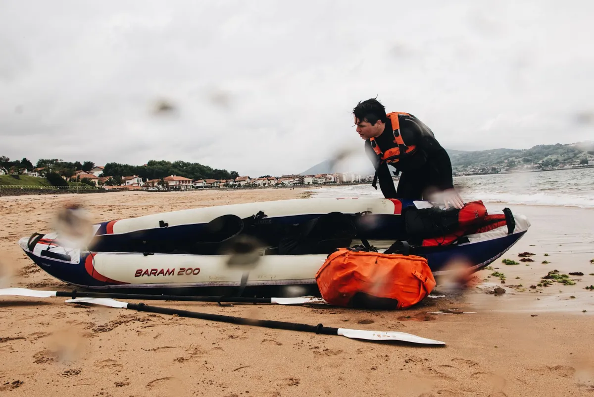 Paddler in a wetsuit beside an inflatable kayak on wet sand with an orange dry bag and paddles