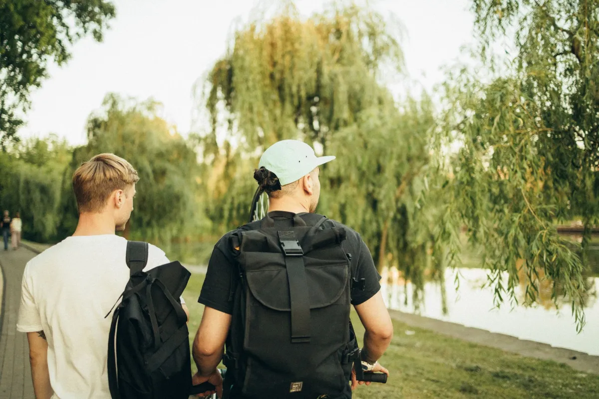 Two people walking a riverside path with roll-top backpacks on their shoulders