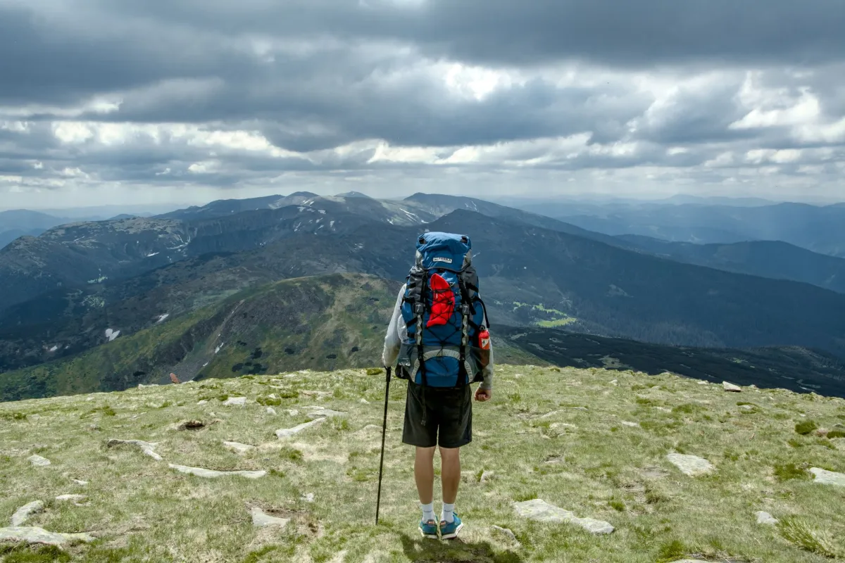 Hiker standing on a grassy ridge with a large lidded expedition backpack and a trekking pole