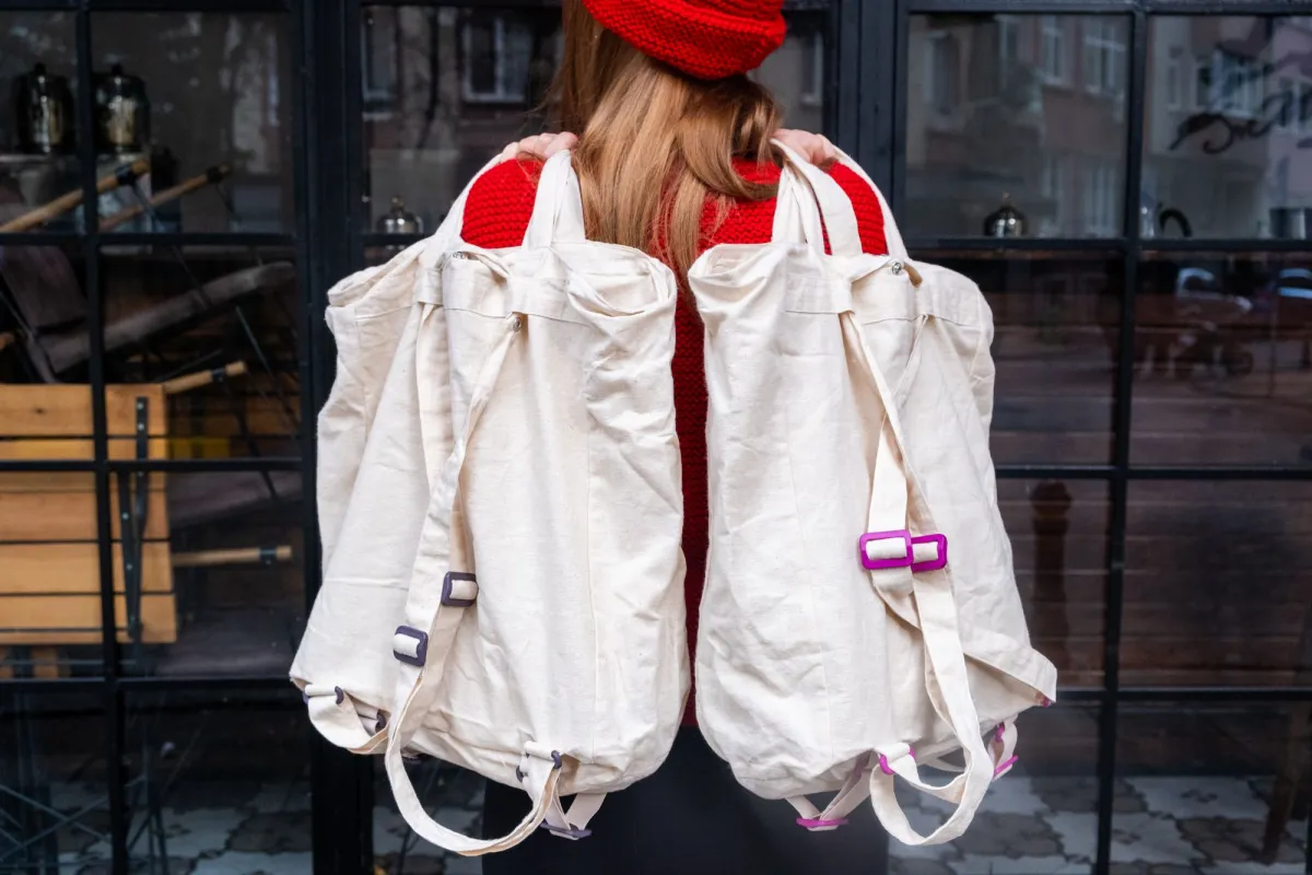 Person in a red hat holding two cream canvas rucksacks with drawstring tops