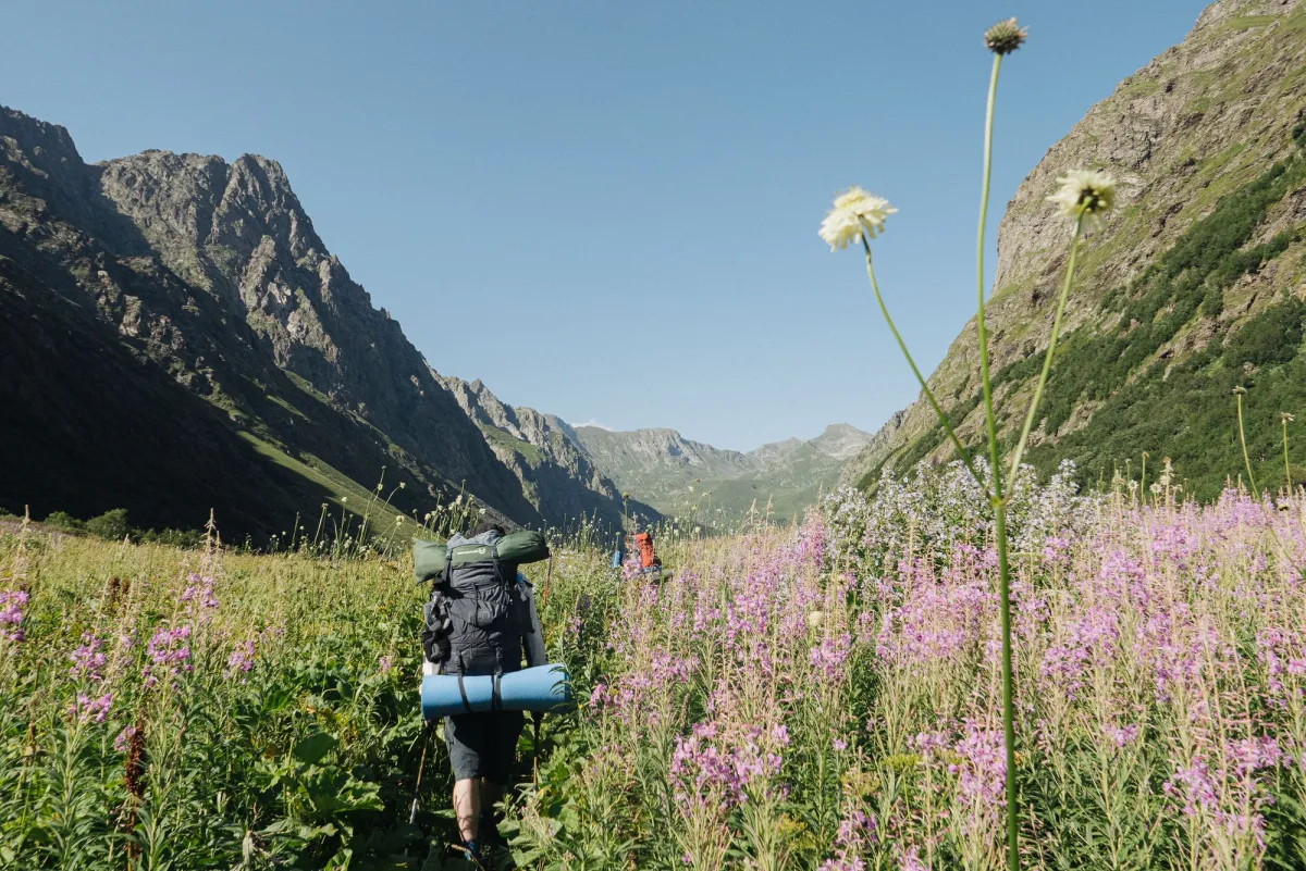 Backpacker walking through a wildflower valley with a lidded pack and a sleeping mat strapped underneath