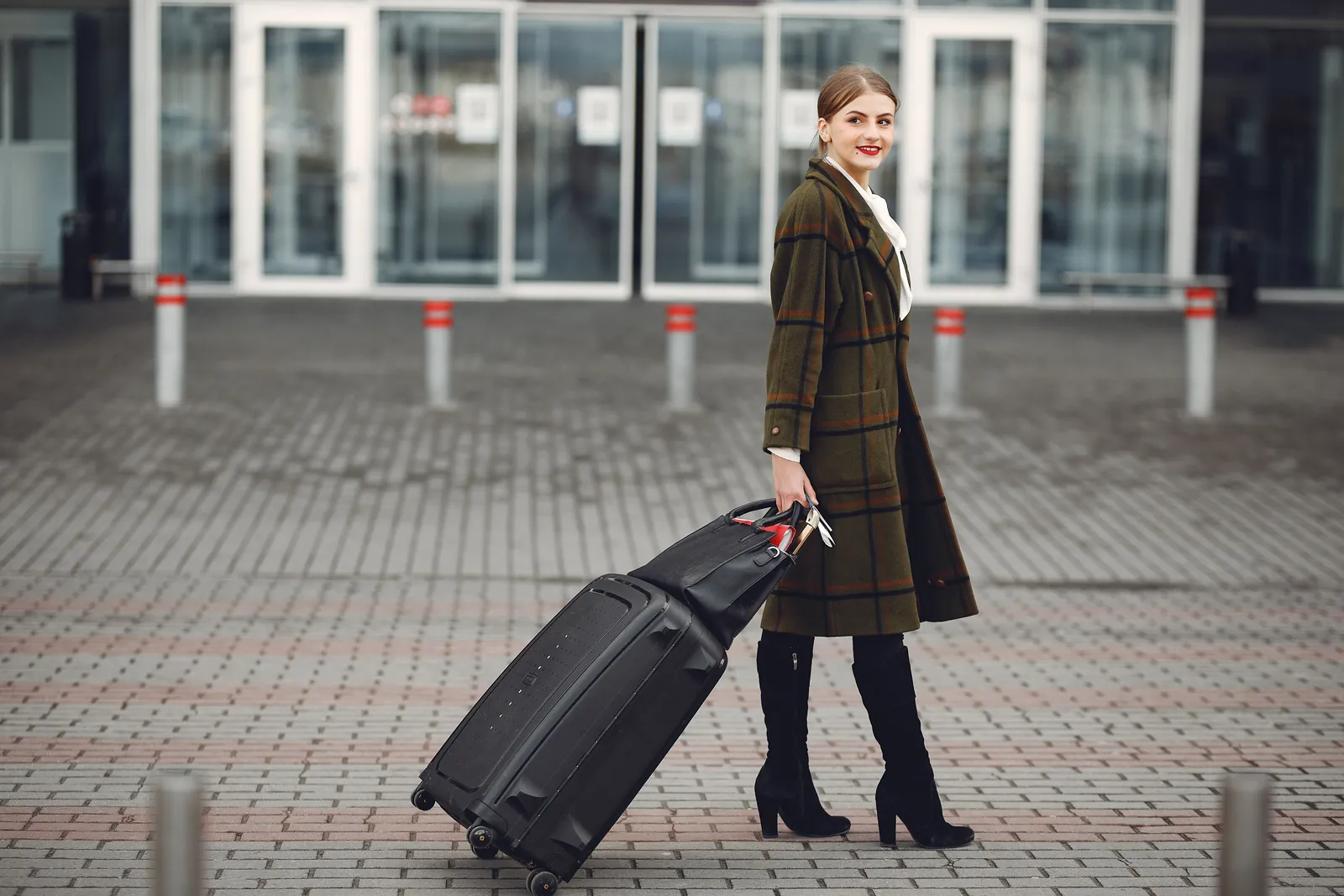 A traveler in a long plaid coat pulls a black spinner suitcase on its wheels across the paved forecourt of an airport terminal