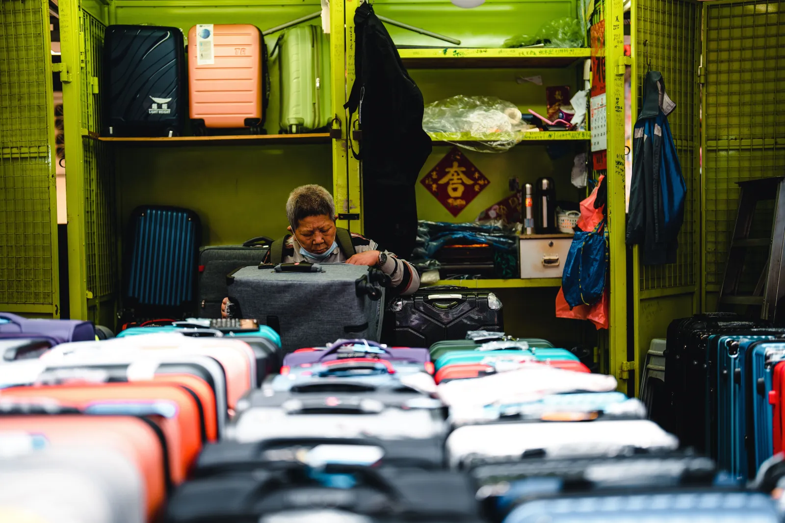 A shopkeeper checks a gray suitcase behind rows of hardside luggage in a small market shop, with more suitcases stacked on the shelves