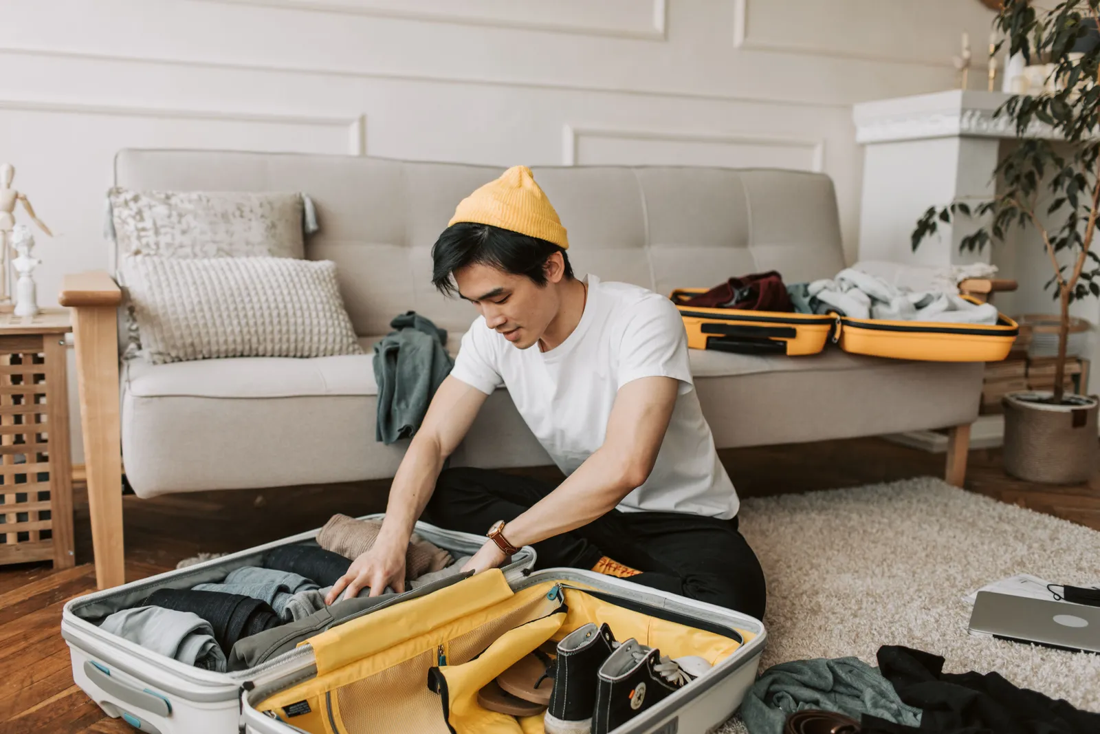 A young man in a yellow beanie kneels on a living room floor packing clothes into an open white hardside suitcase with a yellow lining