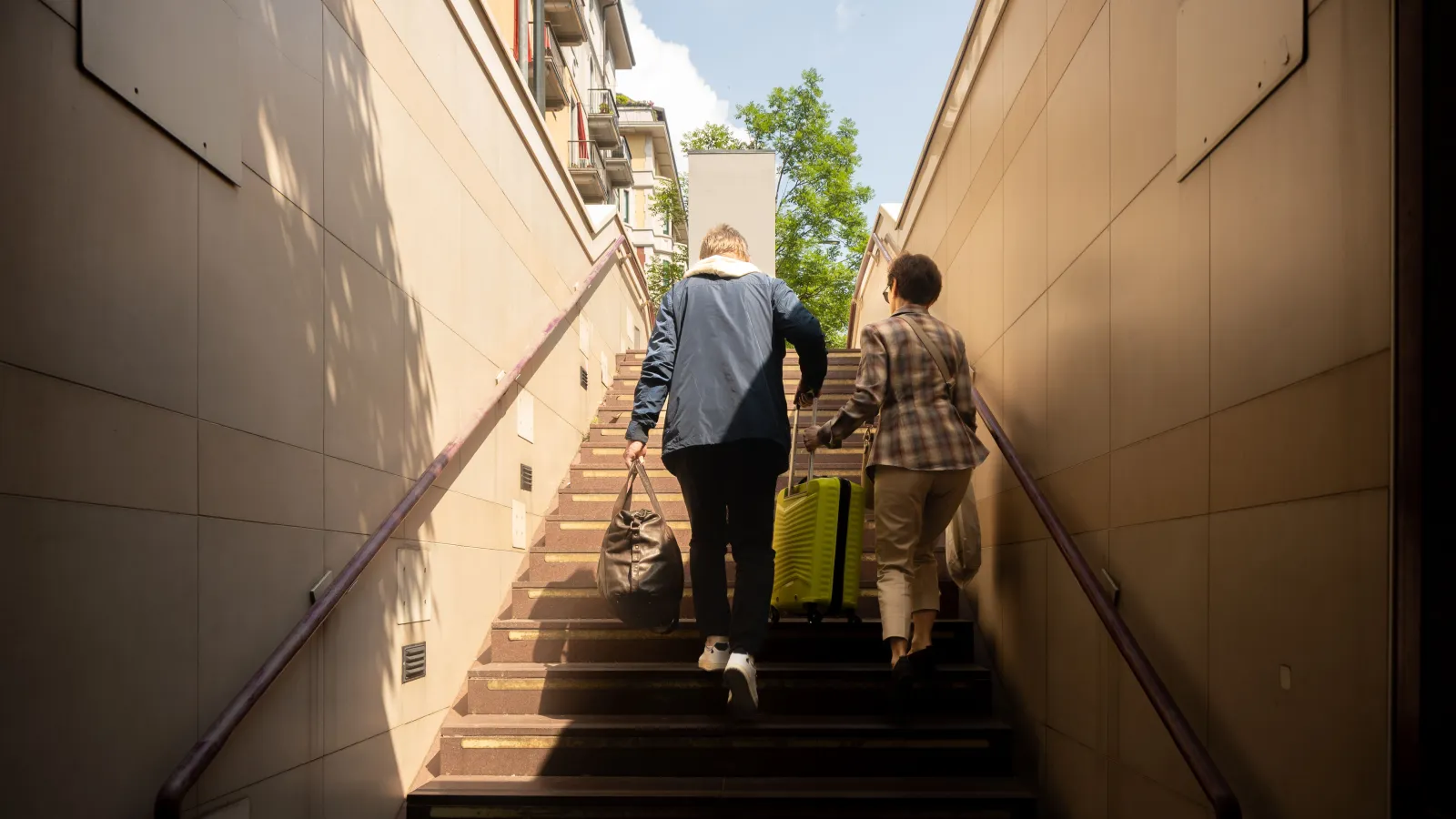 Two travelers climbing a sunlit outdoor staircase, one carrying a dark leather duffel and pulling a lime green suitcase
