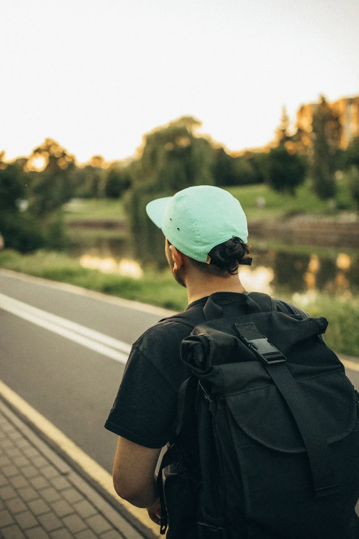 Person in a cap with a black backpack walking a lakeside road at sunset