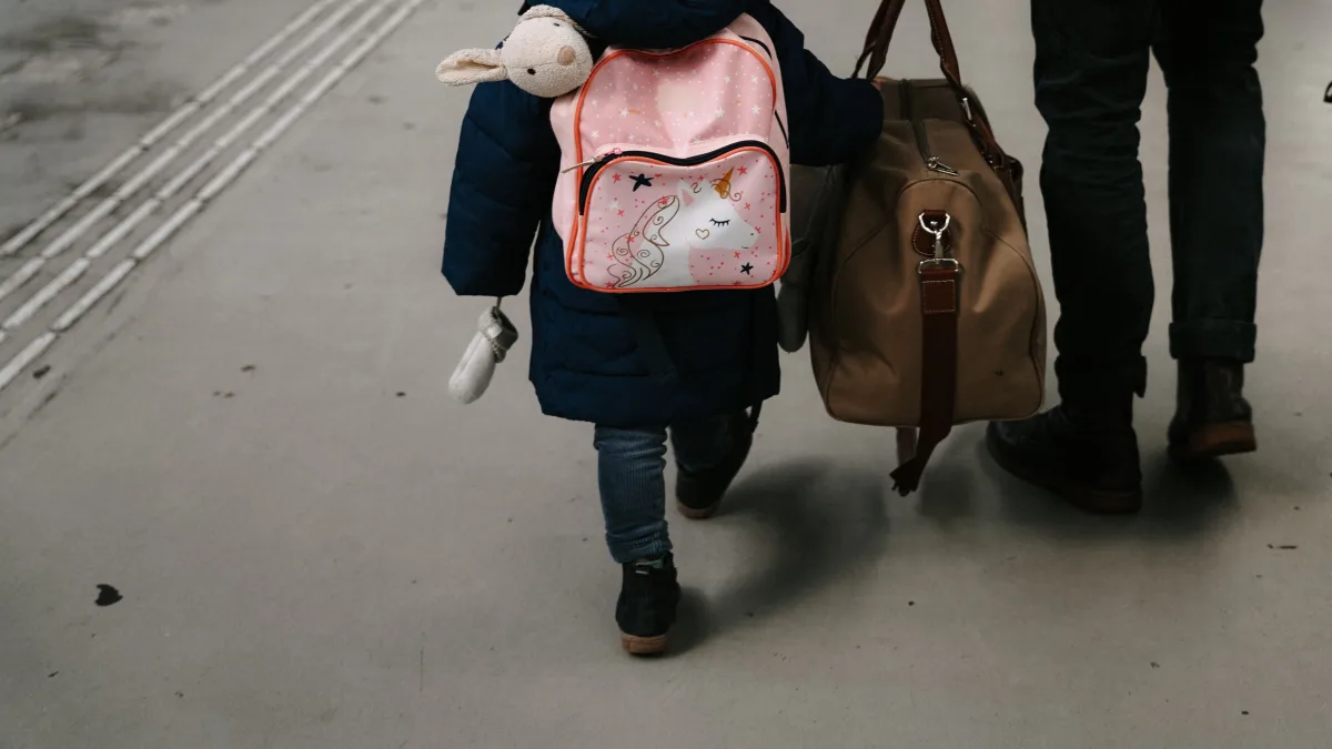 A small child in a winter coat and pom-pom hat, seen from behind, wearing a pink unicorn backpack and walking beside an adult carrying a tan duffel on a station platform