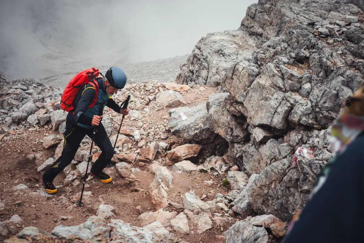 Hiker in a helmet with a red pack climbing a rocky scree slope with trekking poles