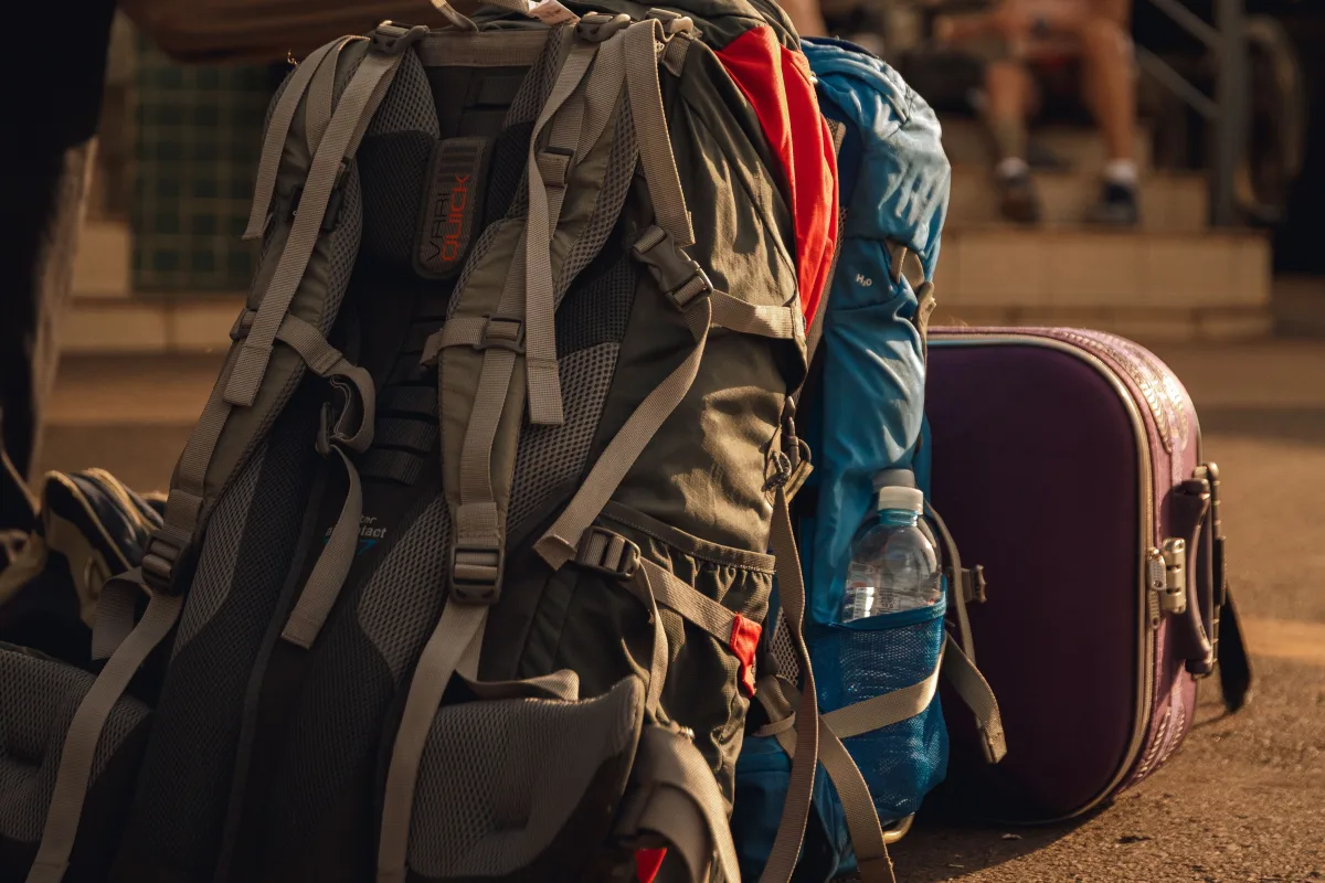 Two hiking backpacks with loose shoulder straps and buckles hanging, set on the ground beside a purple suitcase