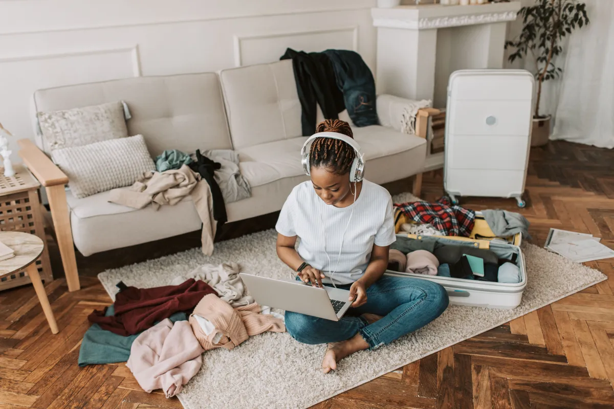 Woman using a laptop beside an open suitcase and clothes on the floor.