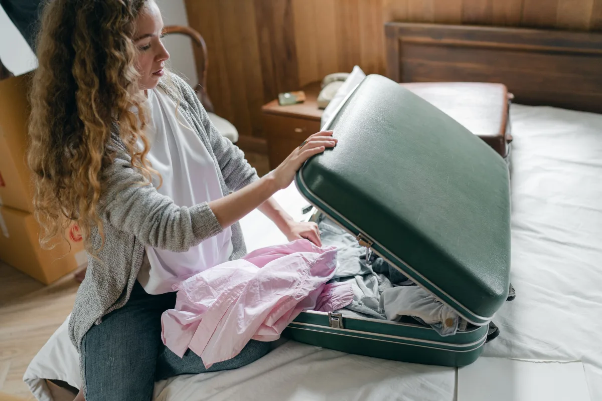 Woman kneeling on a bed, lifting the lid of an overstuffed green vintage suitcase full of clothes
