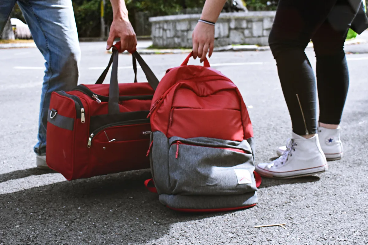 Two travelers lifting a red duffel and a packed red and gray backpack by their grab handles on a street
