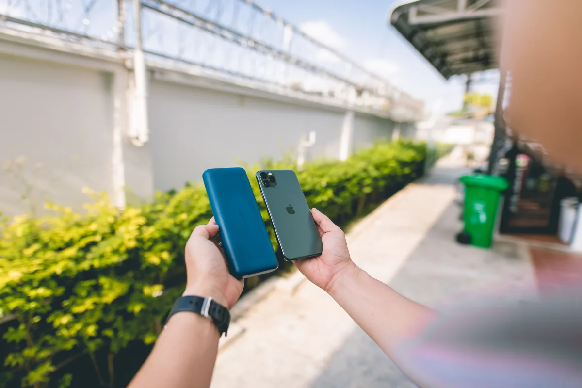 Hands holding a blue power bank and a smartphone outdoors beside a hedge