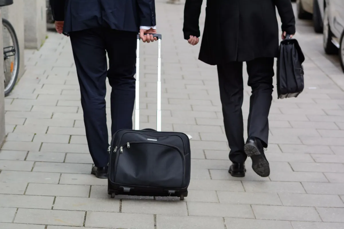 Two business travelers in suits walking down a city sidewalk with a black rolling carry-on case and a leather briefcase